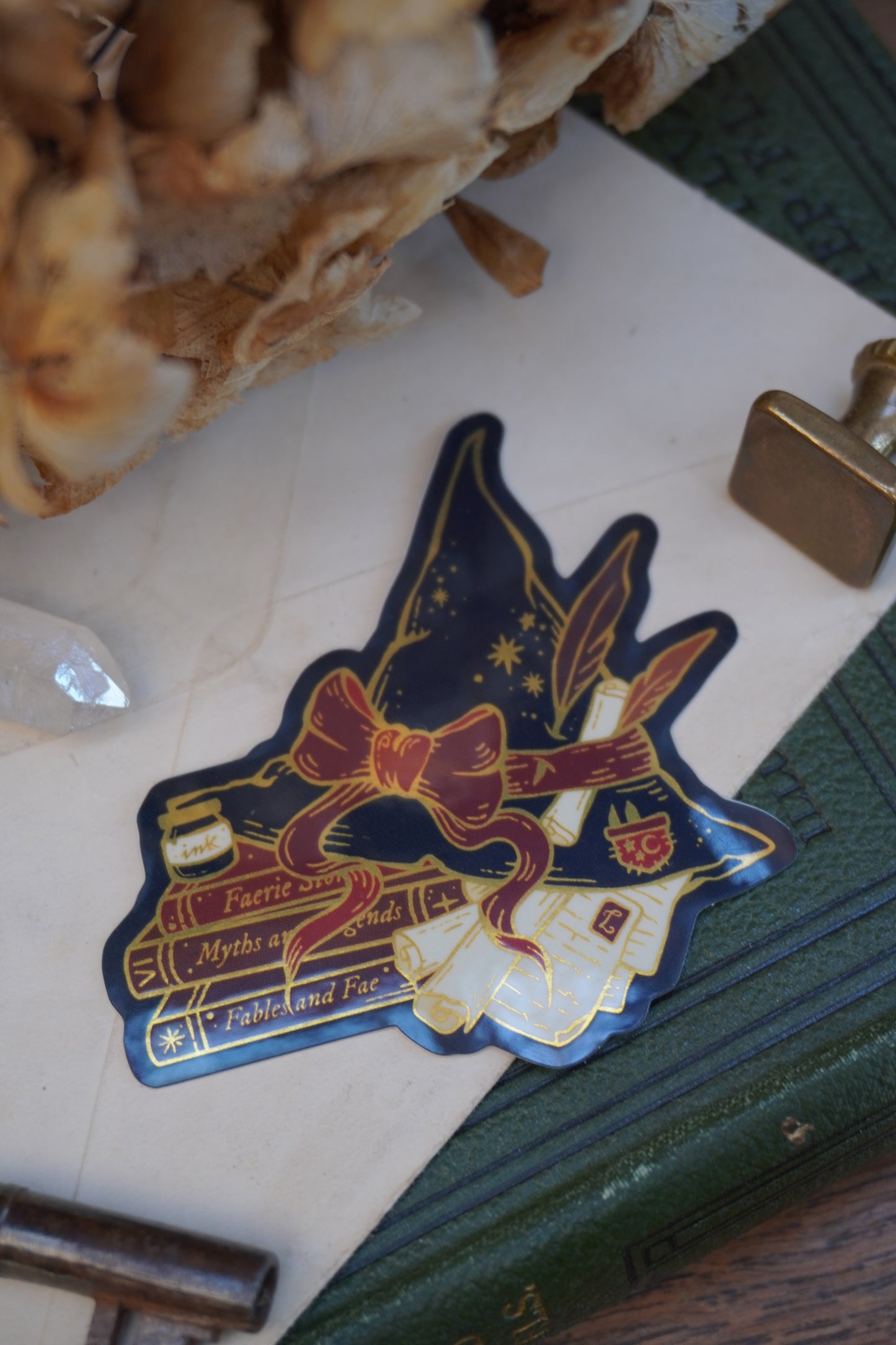 A die-cut sticker shaped into a witch hat on top of books and scrolls, resting on a vintage book next to a brass key, wax seal stamp, crystal and dried flowers.