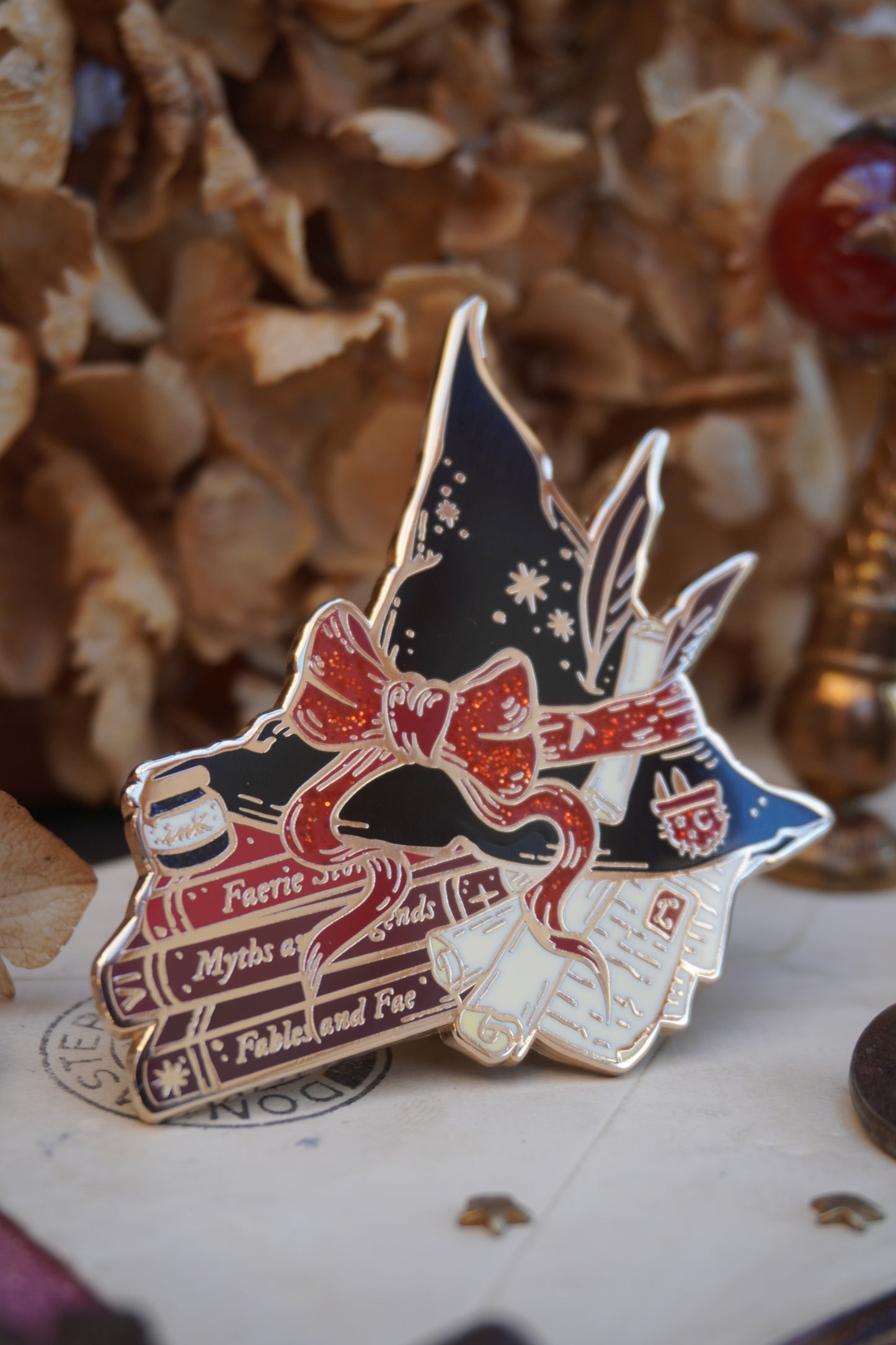 An enamel pin shaped into a witch hat on top of books and scrolls, resting on a vintage style surface with dried flowers in the background.