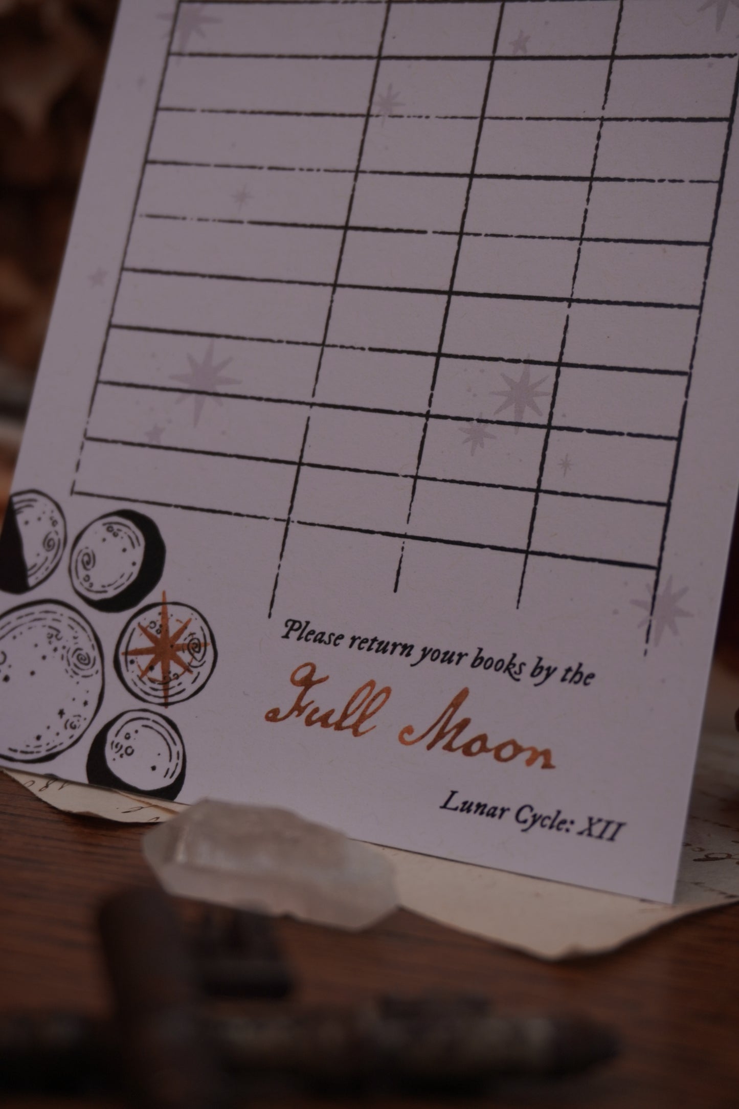 A close-up of a printed library card bookmark with moon phase illustration and the text 'please return your books by the full moon, lunar cycle XII' Photographed on a dark background with dried beige flowers old key and crystal
