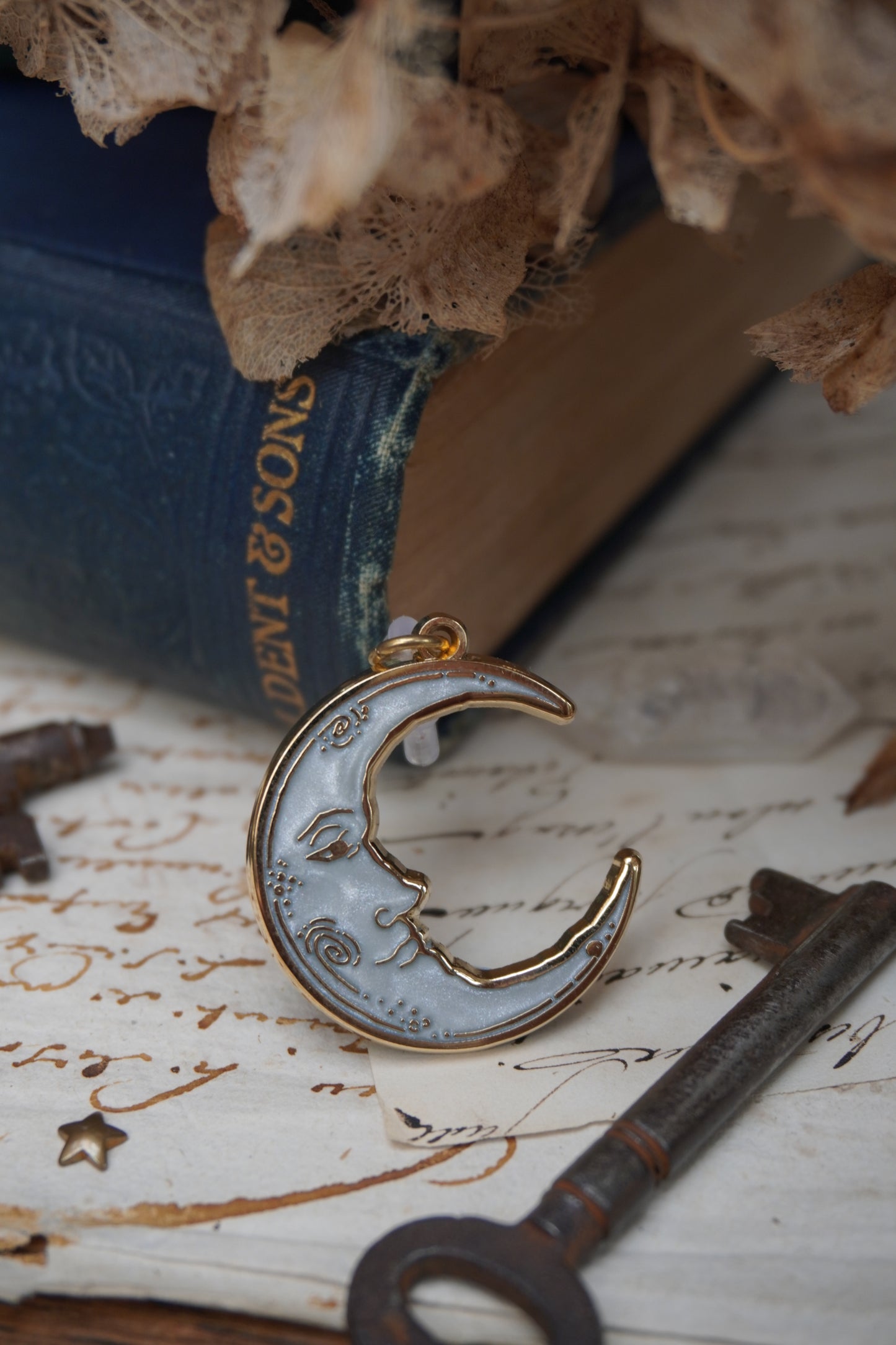 A vintage man in the moon, crescent shaped dust plug with white pearl swirl enamel and gold details rests on vintage paper with a decorative background of vintage books, dried flowers and keys.