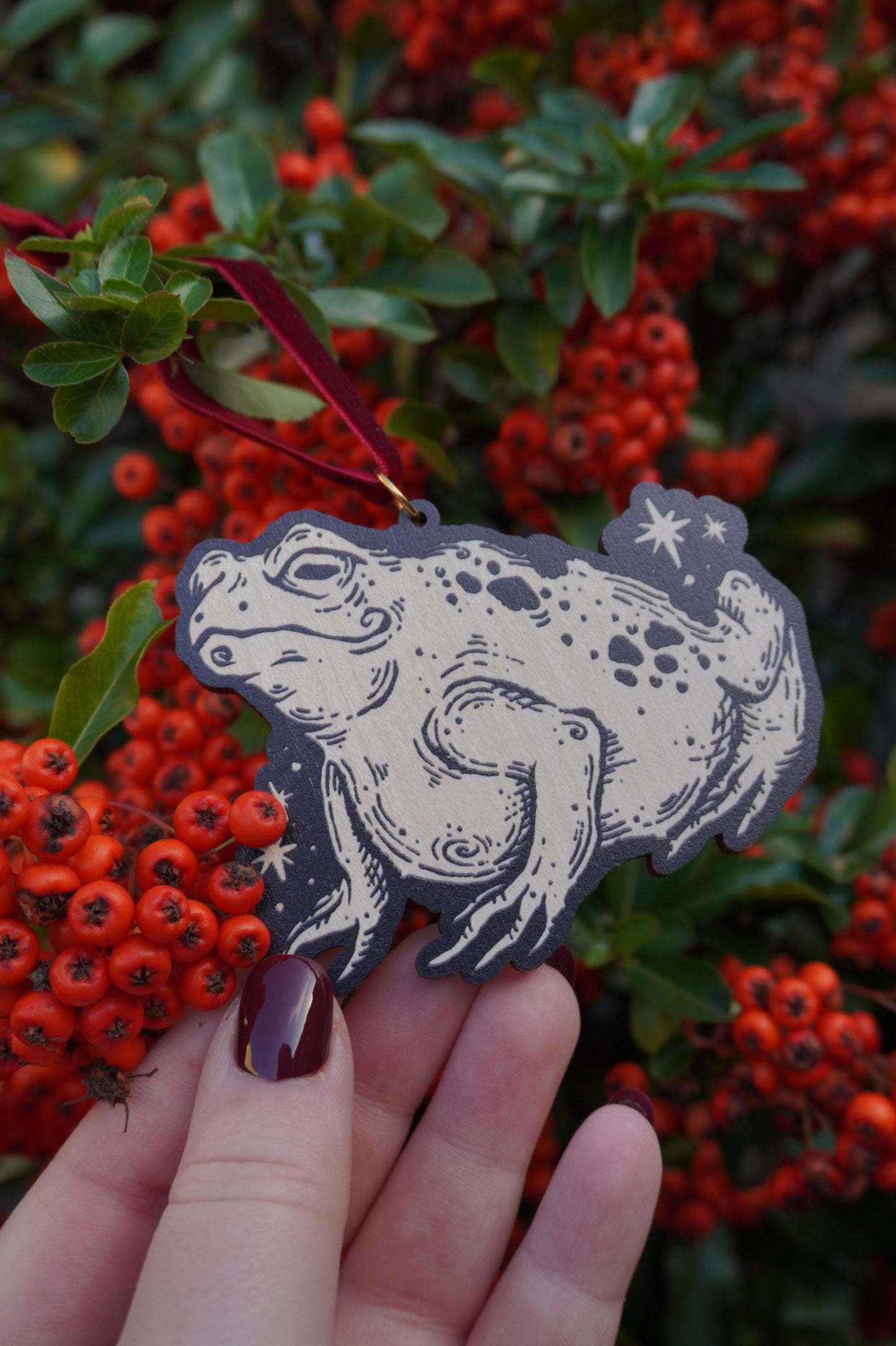 Hand holding a Wooden hanging ornament in the shape of a Toad with black engraving with a red ribbon against a red berry bush