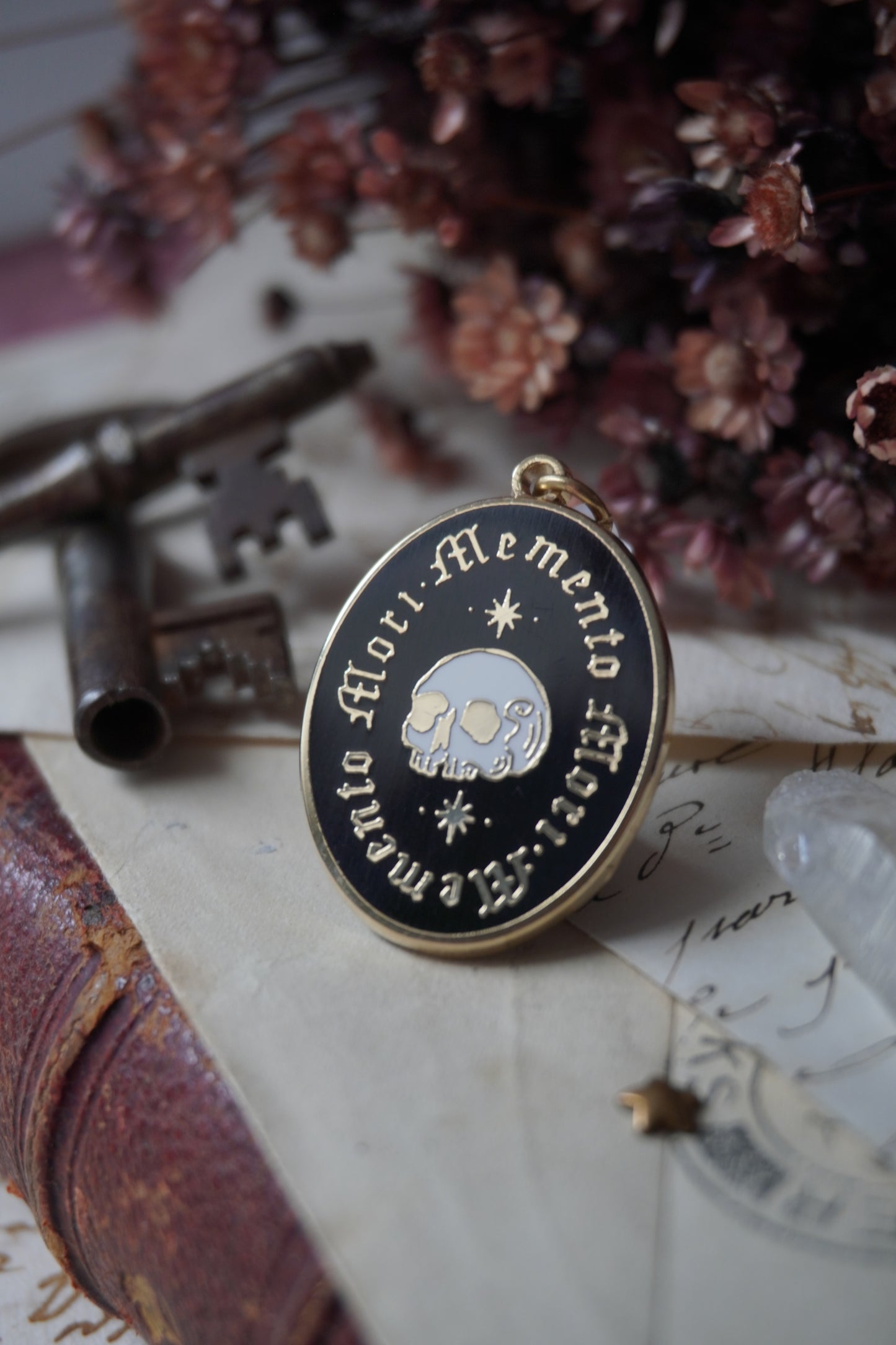 An oval enamel dust plug pendant black background, gold and white skull an stars, and the text 'Memento Mori' framed twice in gold, styled on vintage paper with dried flowers, a vintage key, and a crystal.