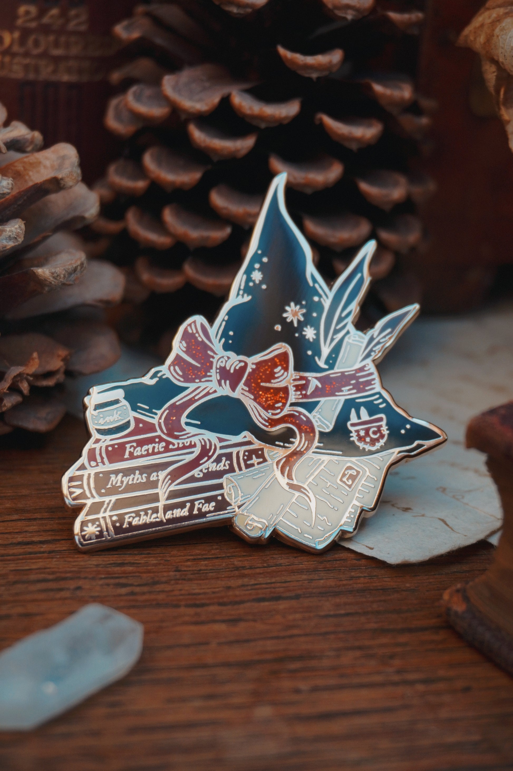 An enamel pin shaped into a witch hat on top of books and scrolls, resting on a wooden surface with decorative elements around like a crystal and pinecone