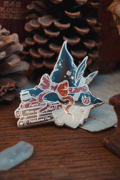 An enamel pin shaped into a witch hat on top of books and scrolls, resting on a wooden surface with decorative elements around like a crystal and pinecone