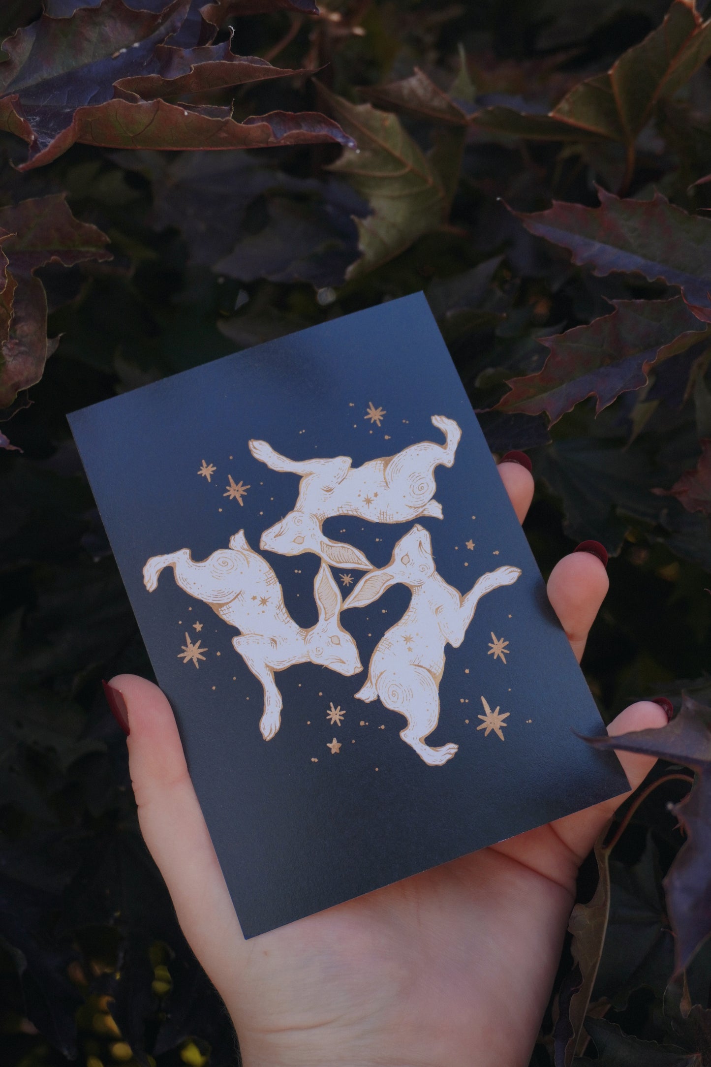 A hand holds a black postcard featuring a white and gold illustration of a triple hare and stars, set against a background of dark purple and green leaves.