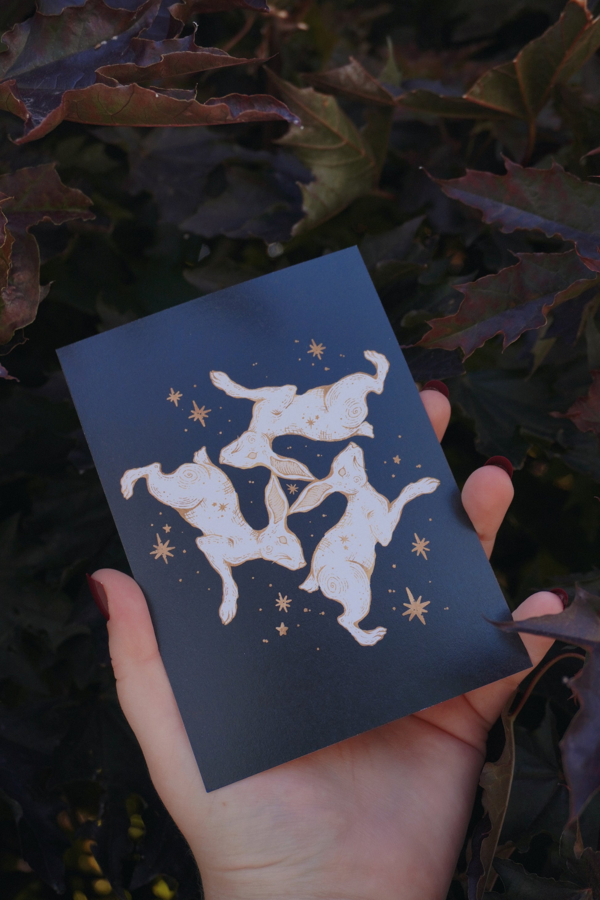 A hand holds a black postcard featuring a white and gold illustration of a triple hare and stars, set against a background of dark purple and green leaves.