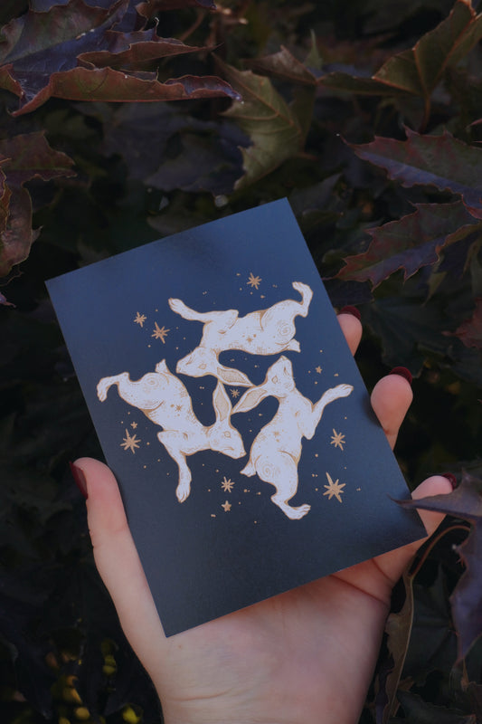 A hand holds a black postcard featuring a white and gold illustration of a triple hare and stars, set against a background of dark purple and green leaves.
