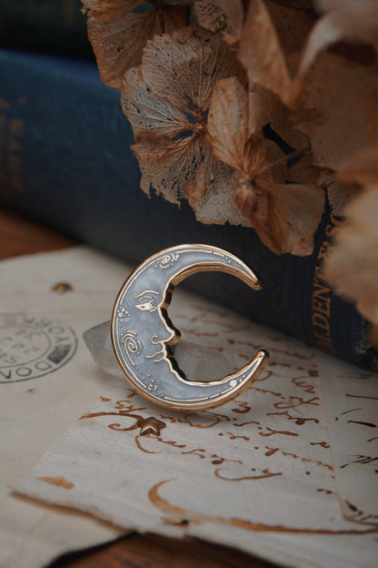 A vintage man in the moon, crescent shaped enamel pin with white pearl swirl enamel and gold details rests on vintage paper with a decorative background of a blue vintage book and dried flowers