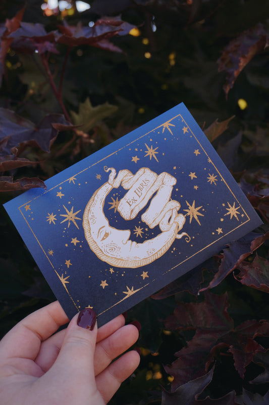 A hand holding a blue toned postcard featuring gold-toned celestial illustration of a crescent moon, stars, and a central banner reading 'Ex Libris'. Set against a background of leaves.