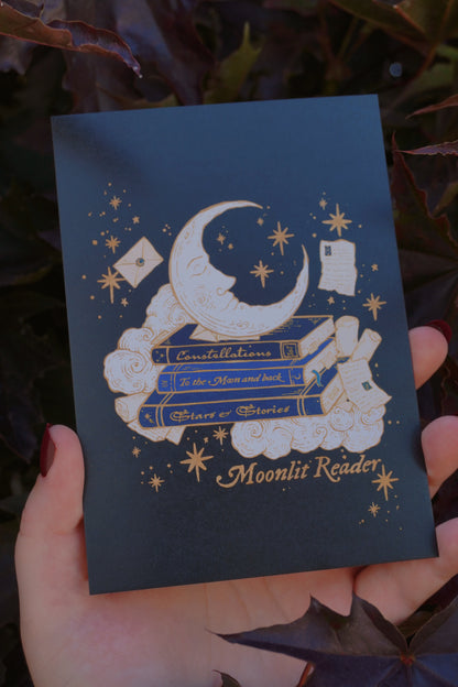 Hand holding a black postcard with a gold white and blue. crescent moon, stars, clouds, books, and scrolls decoration with the text  'Moonlit Reader' aginst dark purple leaves