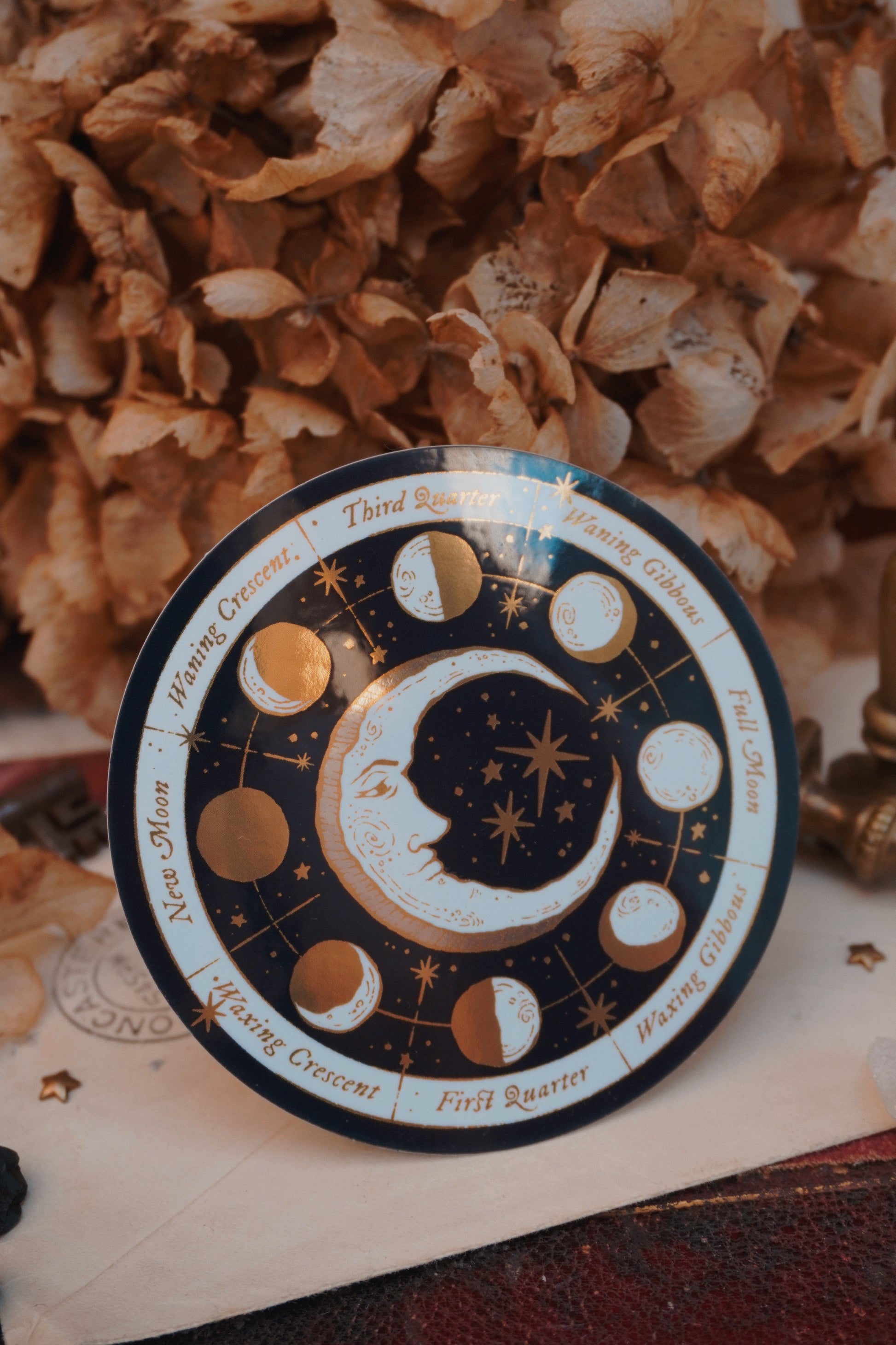 A circular moon phase calendar sticker with gold, cream and white celestial illustrations, and gold text labels, styled on a vintage book with flowers in the background.