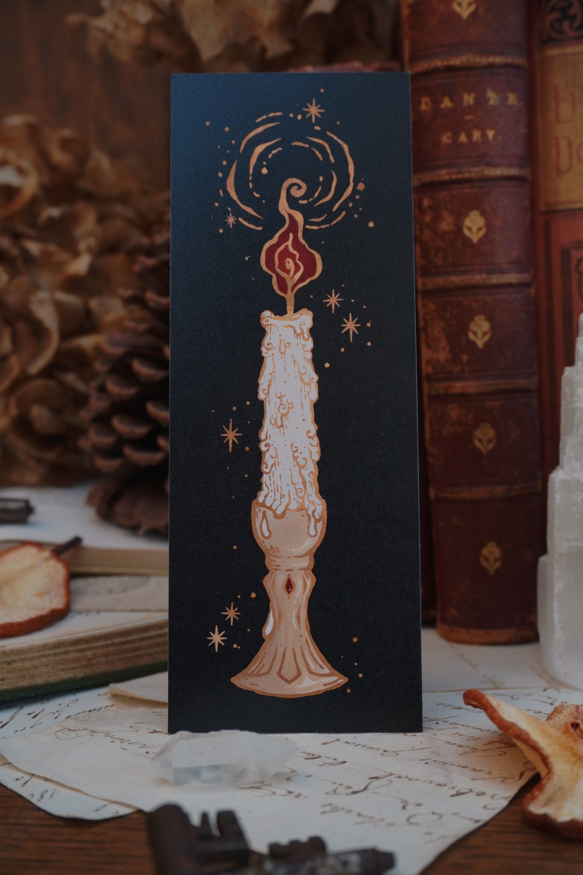 A black bookmark featuring a melting taper candle on a brass holder with stars and a red flame. Outlined with golden hues elements on a vintage style decorative background