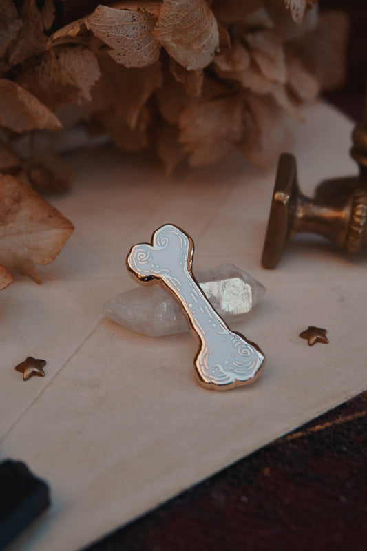 A bone shaped enamel pin in white and gold, resting on a small white crystal displayed on vintage paper on an old book with dried flowers and wax seals in the background