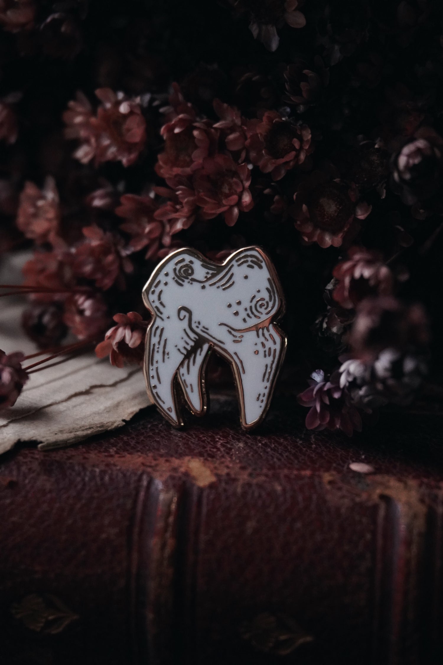 An enamel pin shaped like a tooth with gold plating and white enamel, displayed against a floral background with red vintage book underneath