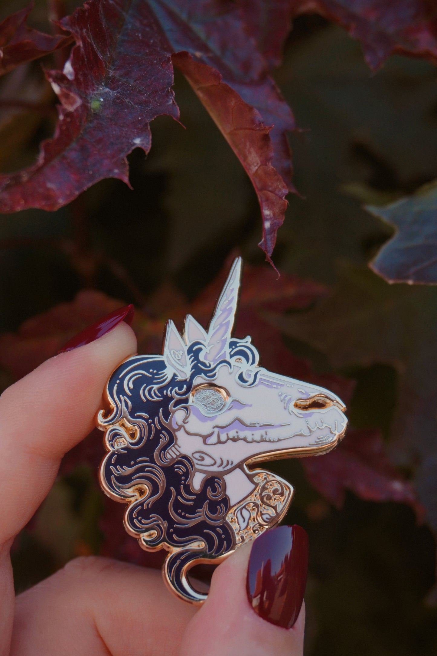 A hand holding a unicorn head enamel pin. The pin features a pink face and neck with white skull on top of the face, dark purple curly mane, lighter purple screen print details and gold plated metal outlines, set against a background of dark red leaves