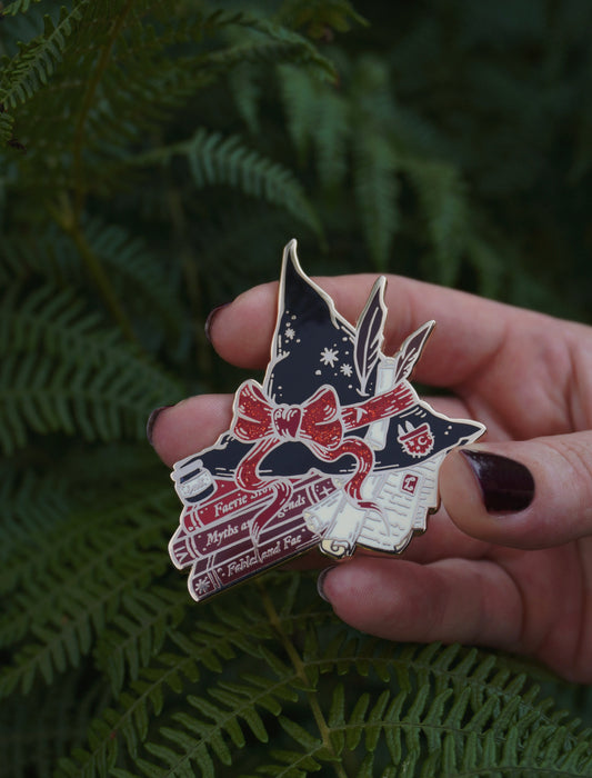 A hand with dark nail polish holds a gold plated enamel pin shaped like a witch's hat on top of books/scrolls, decorated with red, black and white details, against a background of green fern leaves.