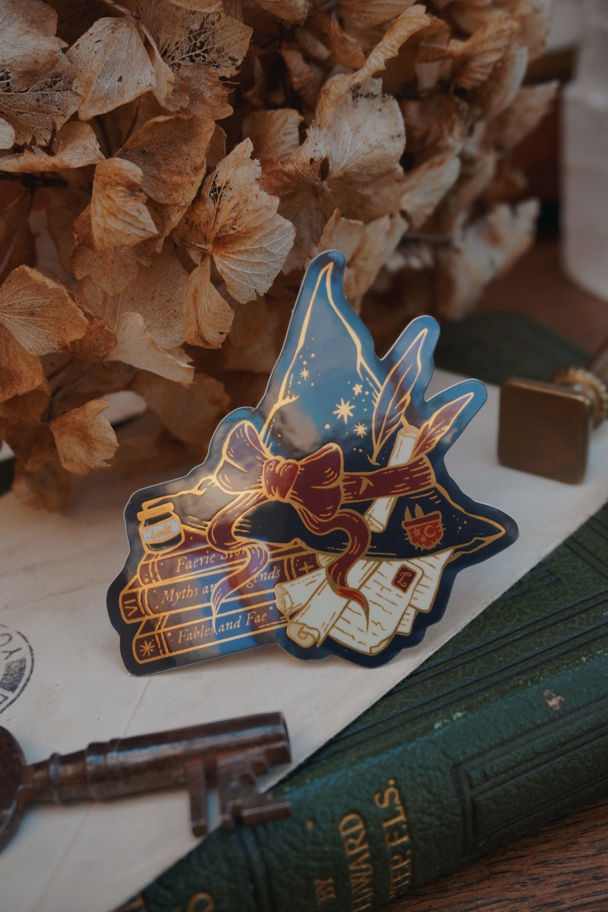 A die-cut sticker shaped into a witch hat on top of books and scrolls, resting on a vintage green book next to a brass key and dried flowers.