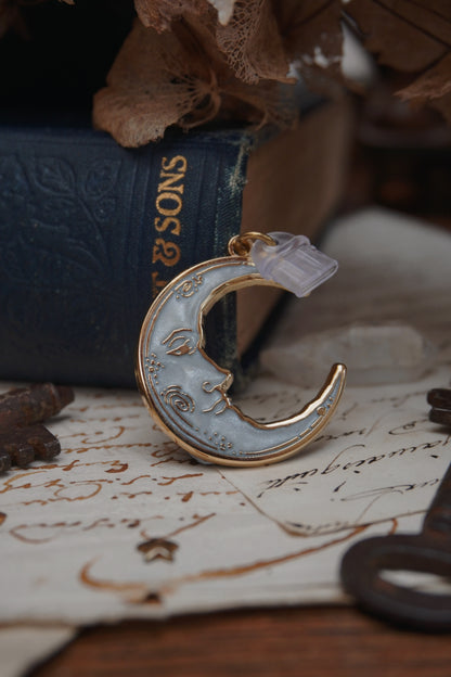 A vintage man in the moon, crescent shaped dust plug with white pearl swirl enamel and gold details rests on vintage paper with a decorative background of vintage books, dried flowers and keys.