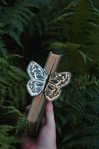 A hand holds a vintage book with a 3ddecorative black and gold foil butterfly bookmark peeking out of the pages set against green ferns