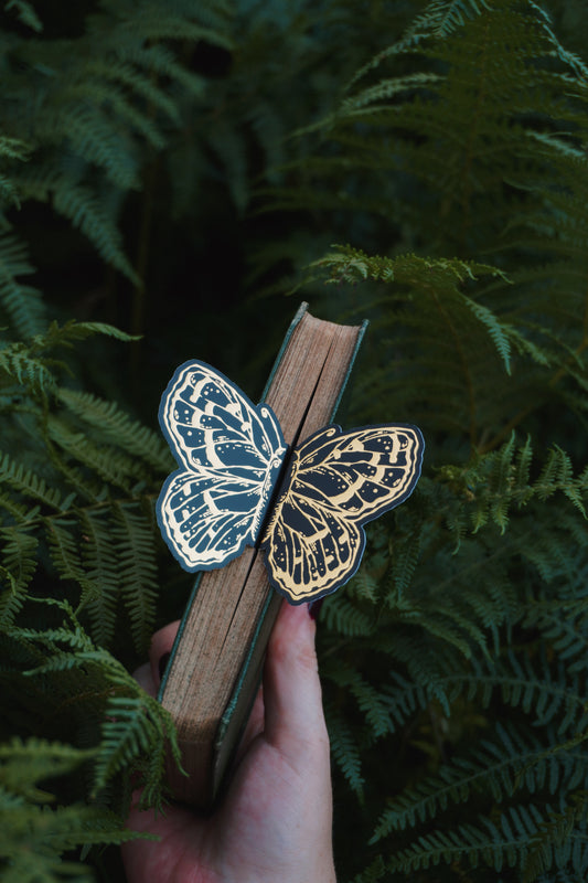 A hand holds a vintage book with a 3ddecorative black and gold foil butterfly bookmark peeking out of the pages set against green ferns