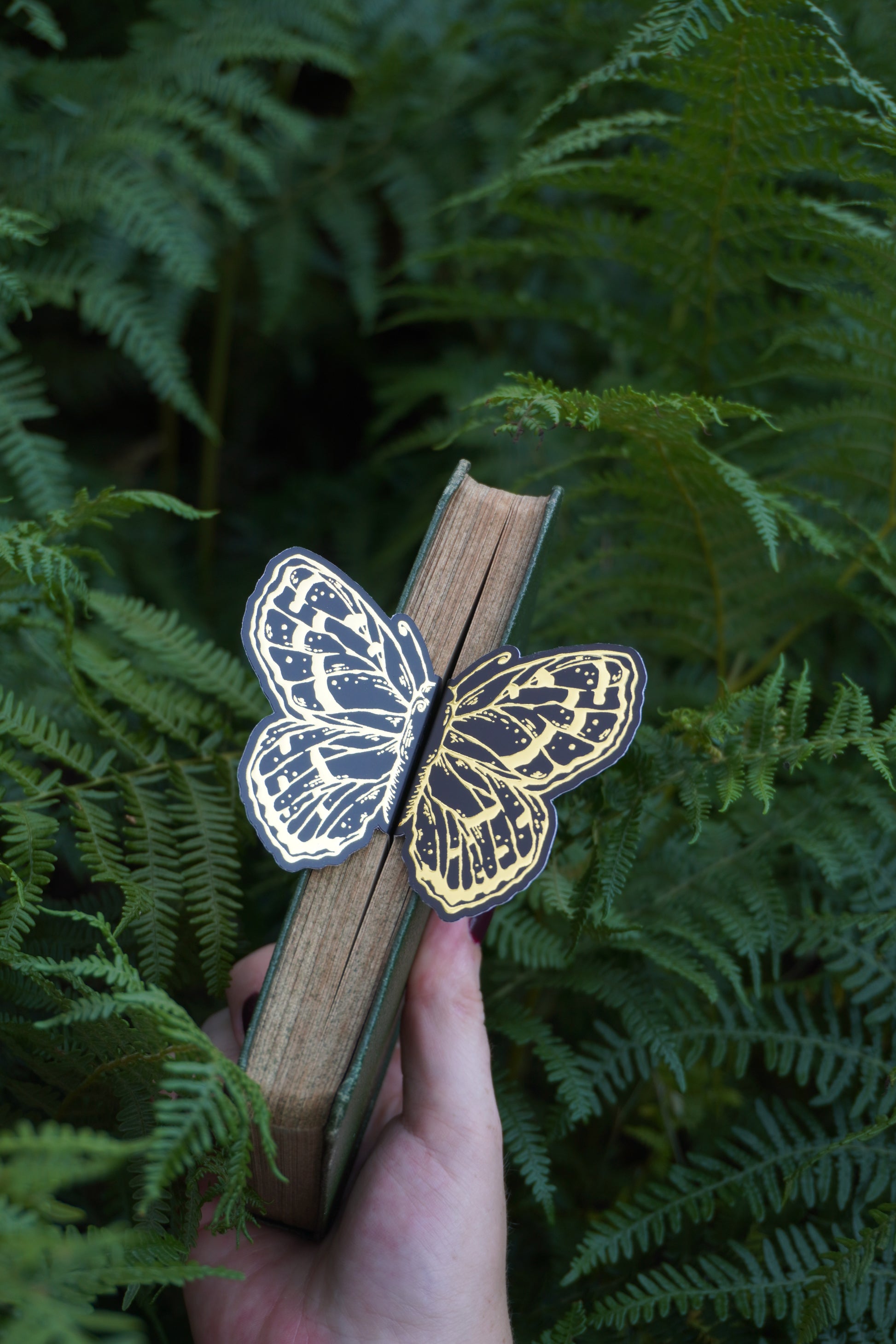 A hand holds a vintage book with a 3d decorative black and gold foil butterfly bookmark peeking out of the pages set against green ferns