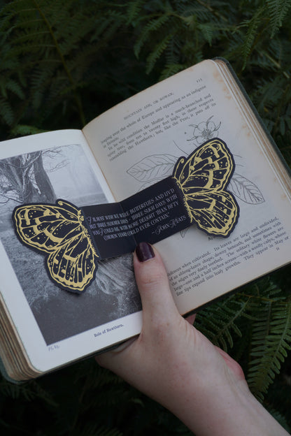 A black paper and gold foil bookmark with gold butterfly illustrations and a white text quote by John Keats ''I almost wish we were butterflies and liv'd but three summer days - three such days with you I could fill with more delight than fifty common years could ever contain' - John Keats, placed across an open book set against green ferns