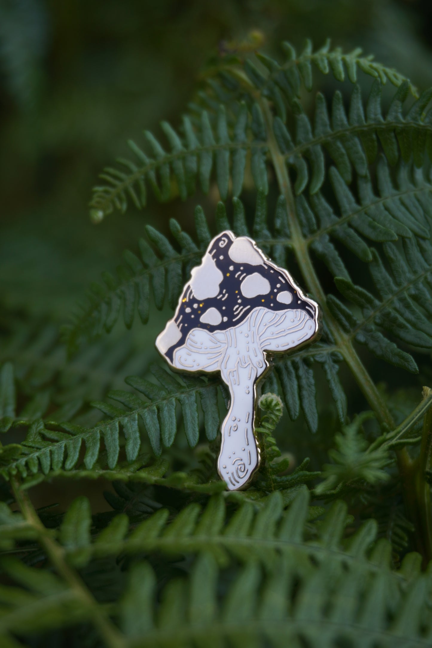 A mushroom shaped enamel pin with a black and gold spotted cap with gold glitter flecks and white stem, photographed resting on green fern leaves.