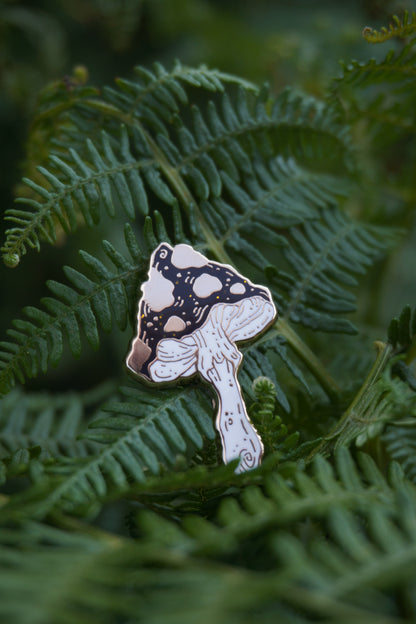 A mushroom shaped enamel pin with a black and gold spotted cap with gold glitter flecks and white stem, photographed resting on green fern leaves.