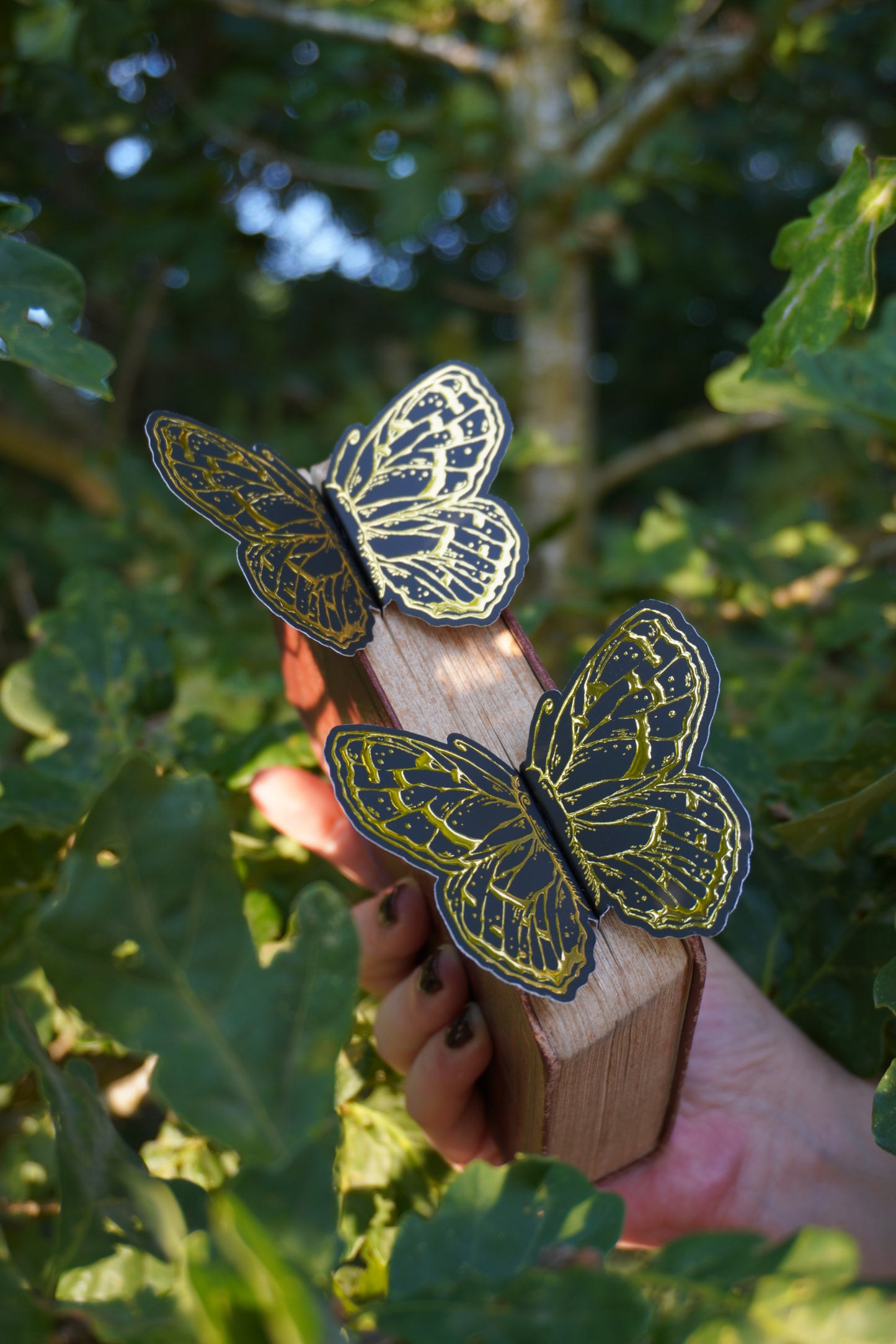 A hand holds a vintage book with two, 3d decorative black and gold foil butterfly bookmark peeking out of the pages set against green oak leaves
