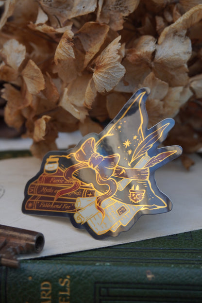 A die-cut sticker shaped into a witch hat on top of books and scrolls, resting on a vintage green book next to a brass key and dried flowers.