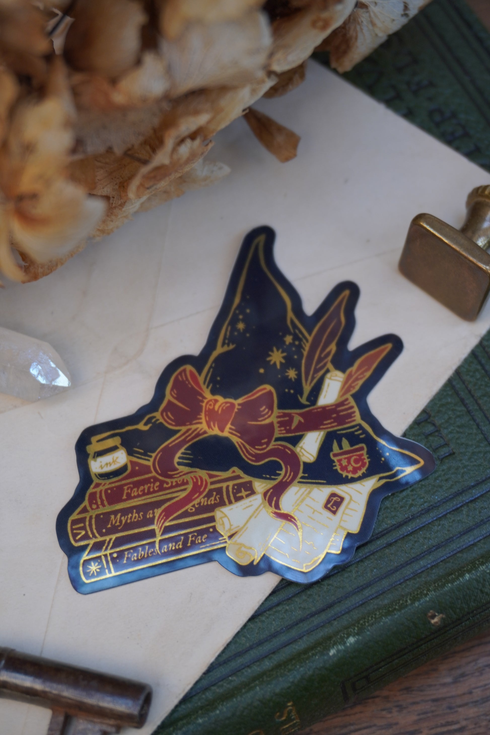 A die-cut sticker shaped into a witch hat on top of books and scrolls, resting on a vintage book next to a brass key, wax seal stamp, crystal and dried flowers.