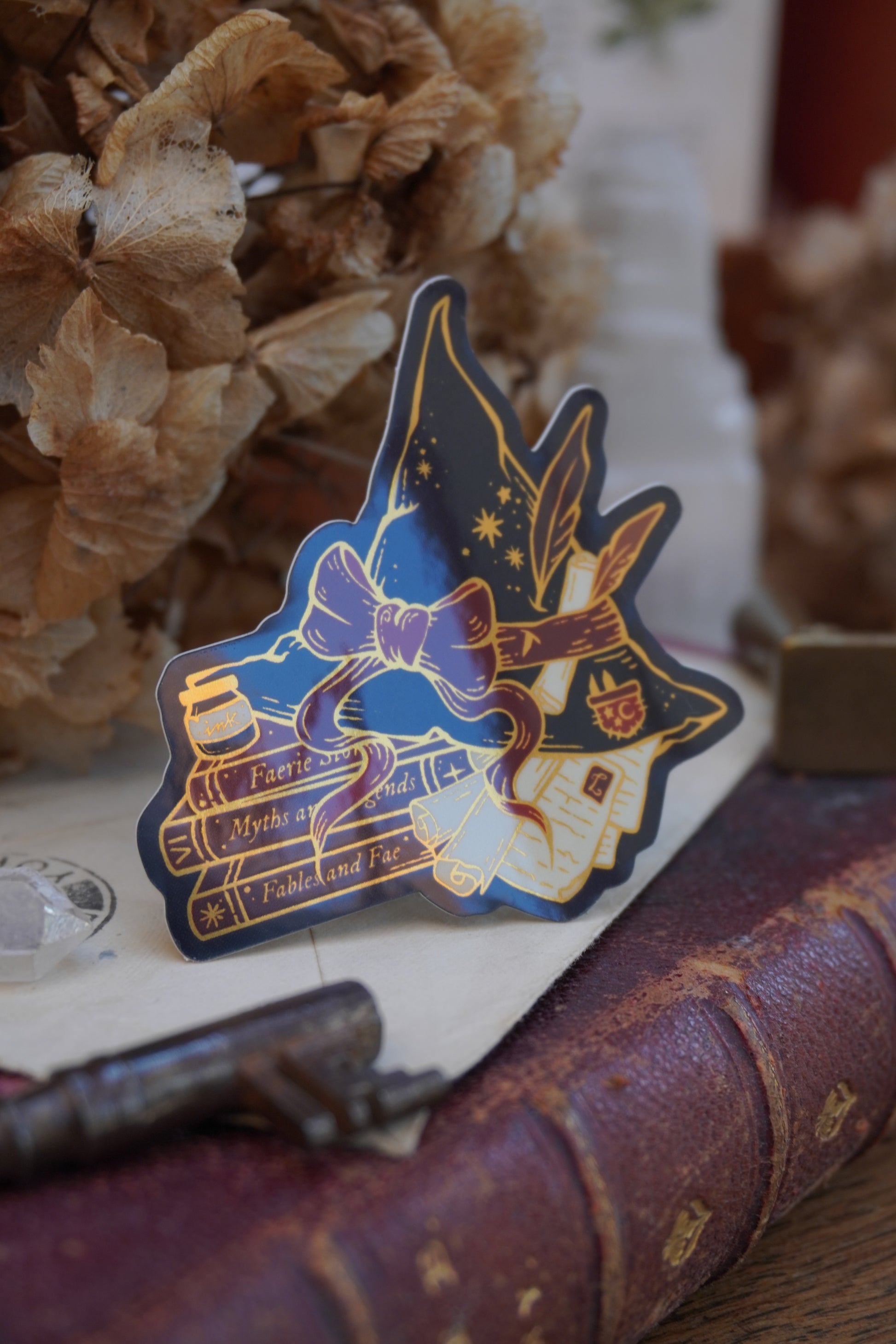 A die-cut sticker shaped into a witch hat on top of books and scrolls, resting on a vintage book next to a brass key, crystal and dried flowers.