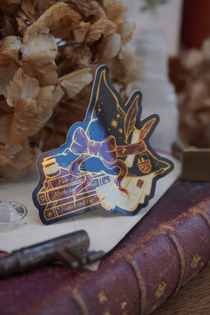 A die-cut sticker shaped into a witch hat on top of books and scrolls, resting on a vintage book next to a brass key, crystal and dried flowers.
