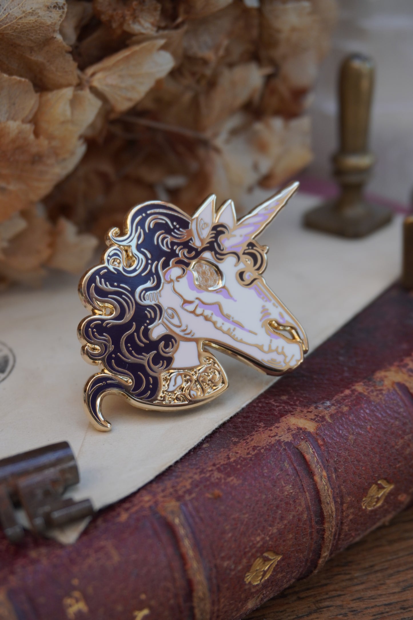 A close-up of an enamel pin shaped like a unicorn head. The pin features a pink face and neck with white skull on top of the face, dark purple curly mane, lighter purple screen print details and gold plated metal outlines resting on vintage paper with dried flowers in the background.