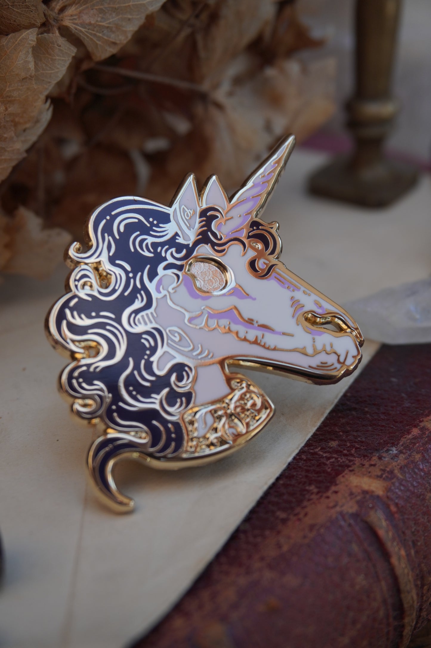 A close-up of an enamel pin shaped like a unicorn head. The pin features a pink face and neck with white skull on top of the face, dark purple curly mane, lighter purple screen print details and gold plated metal outlines resting on vintage paper with dried flowers in the background.