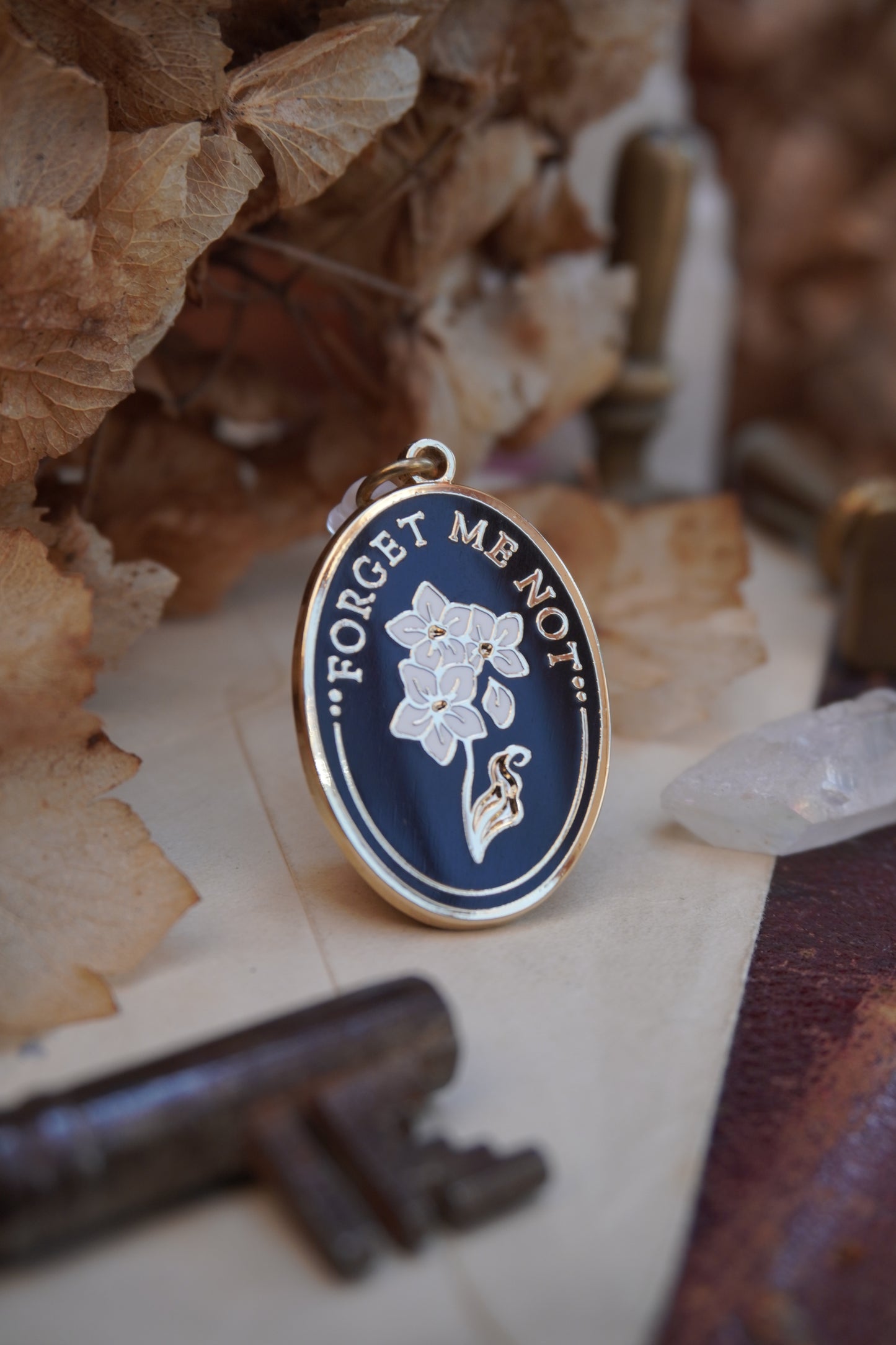 An oval enamel dust plug pendant black background, white floral design, and the text 'FORGET ME NOT' in gold, styled on vintage paper with dried flowers, a vintage key, and a crystal.