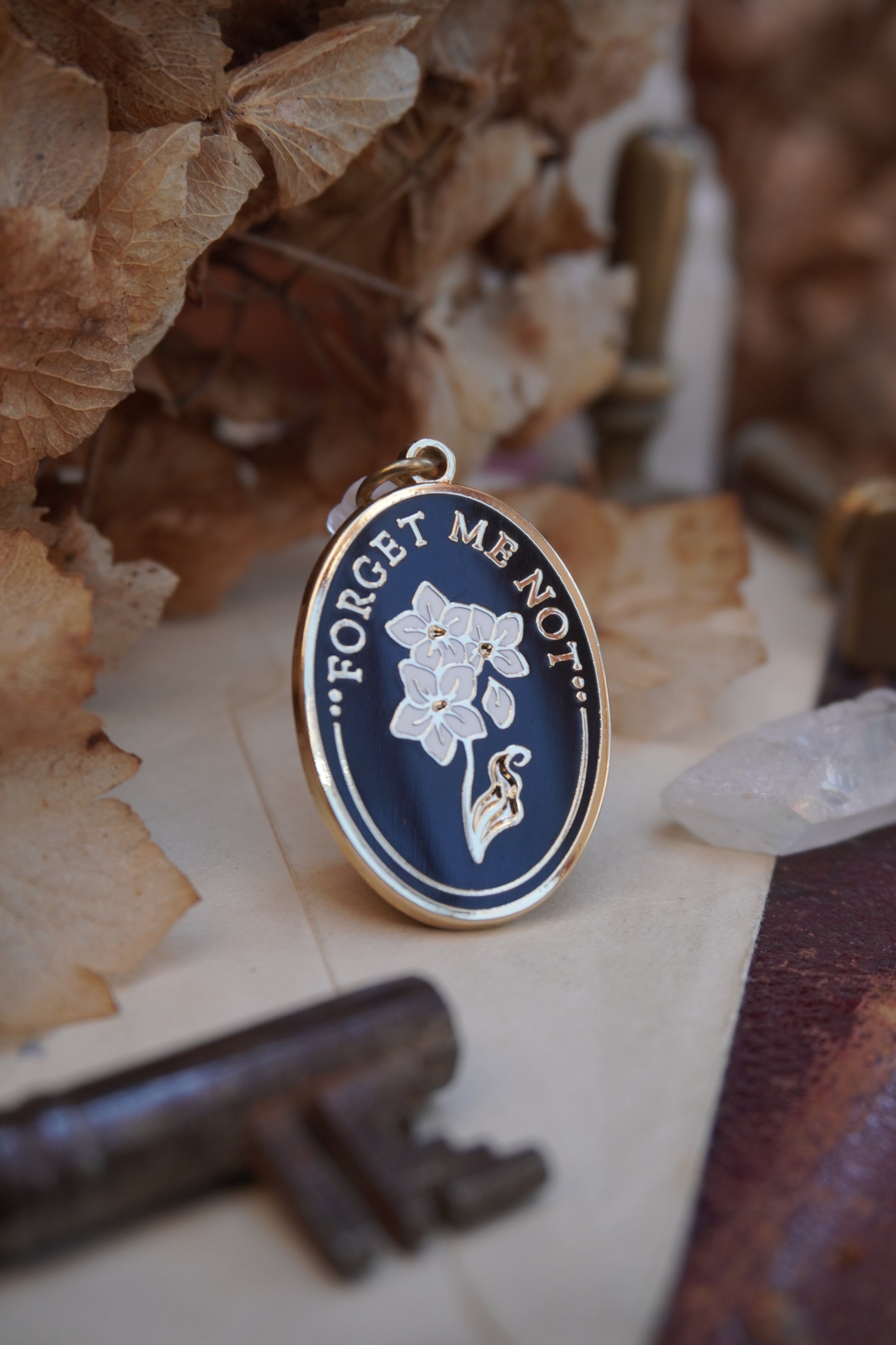 An oval enamel dust plug pendant black background, white floral design, and the text 'FORGET ME NOT' in gold, styled on vintage paper with dried flowers, a vintage key, and a crystal.