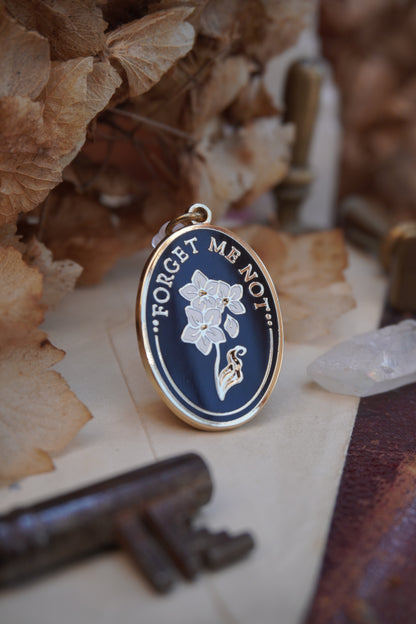 An oval enamel dust plug pendant black background, white floral design, and the text 'FORGET ME NOT' in gold, styled on vintage paper with dried flowers, a vintage key, and a crystal.
