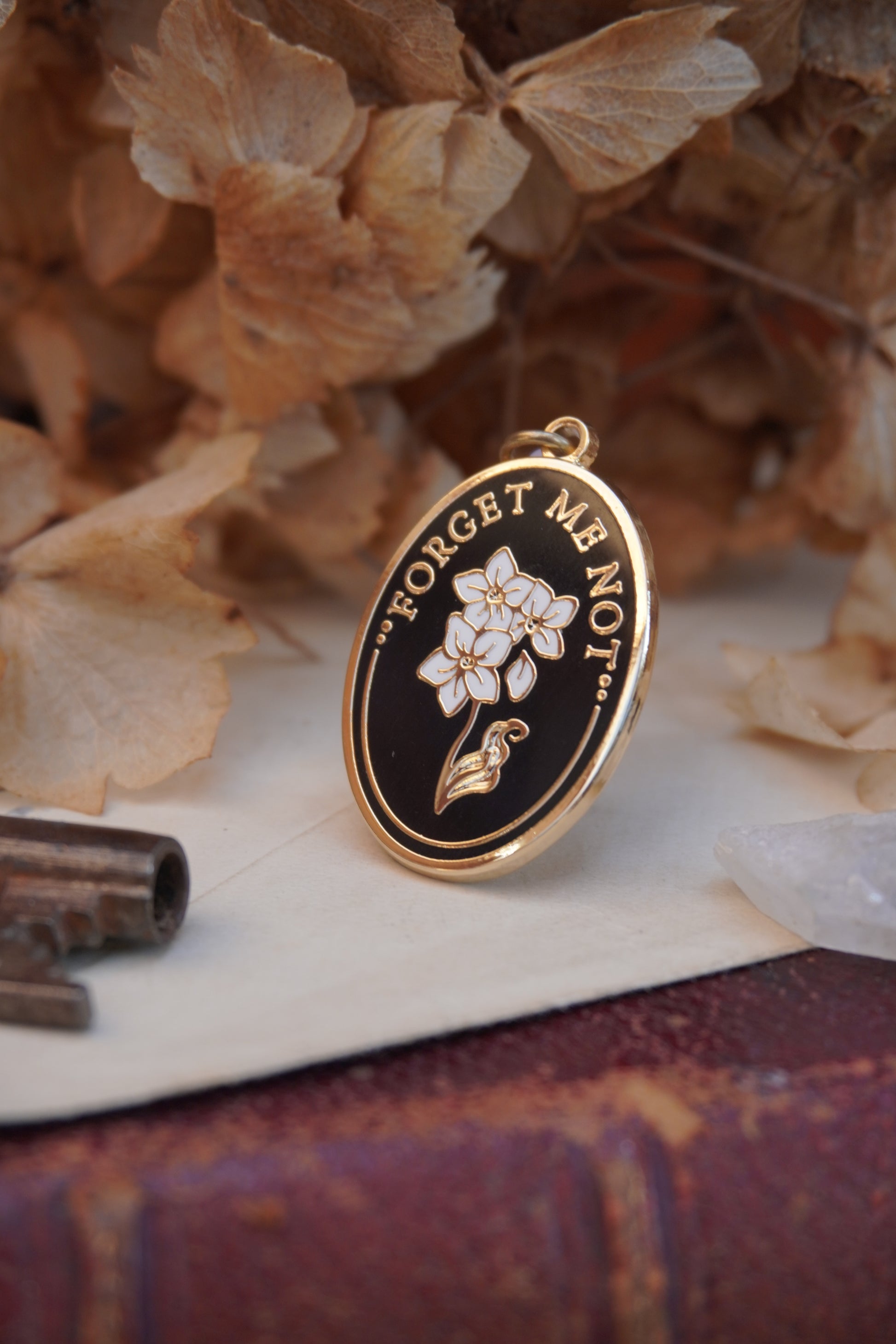An oval enamel dust plug pendant black background, white floral design, and the text 'FORGET ME NOT' in gold, styled on vintage paper with dried flowers, a vintage key, and a crystal.