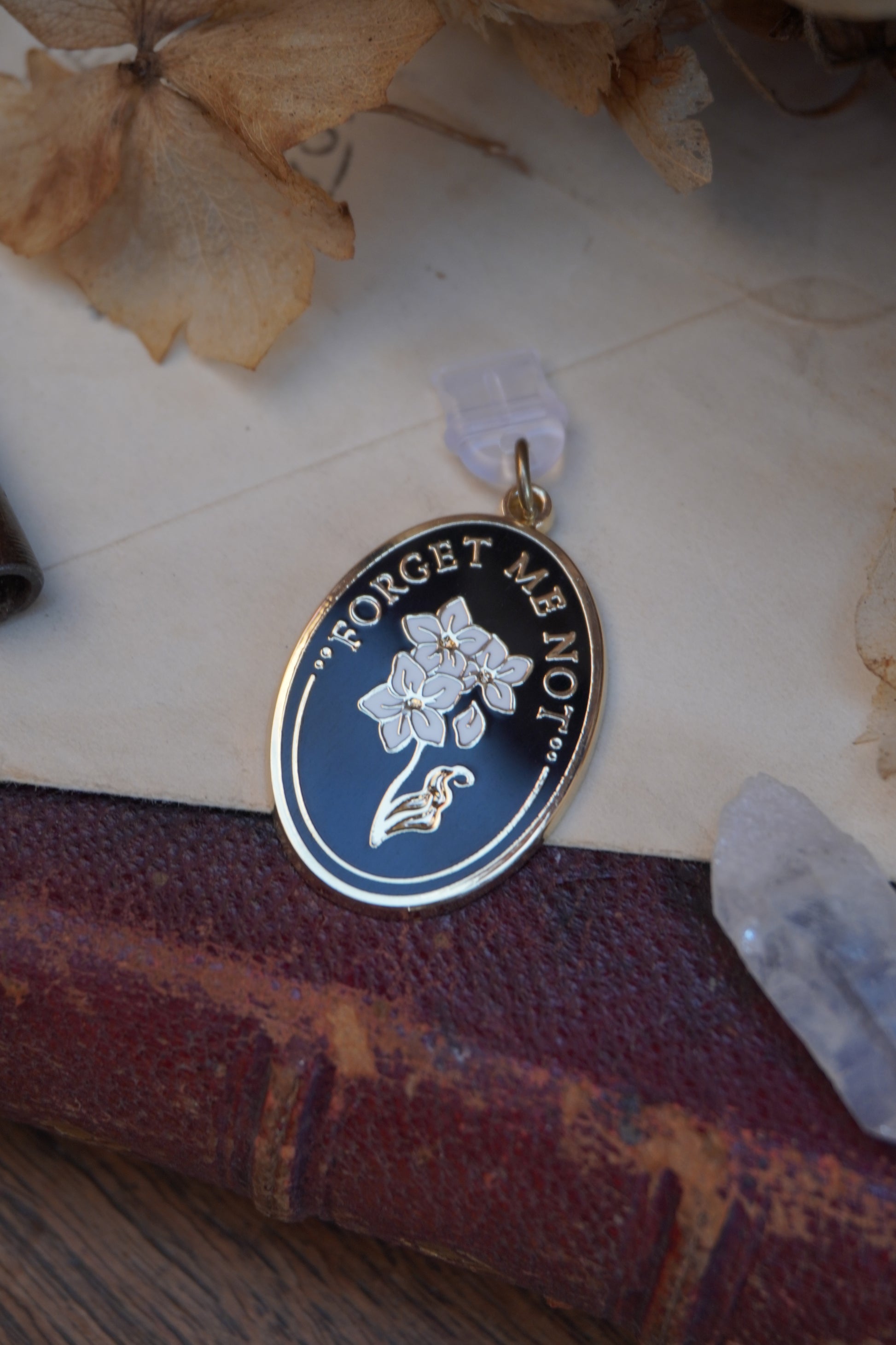 An oval enamel dust plug pendant black background, white floral design, and the text 'FORGET ME NOT' in gold, styled on vintage paper and red book with dried flowers, a vintage key, and a crystal.