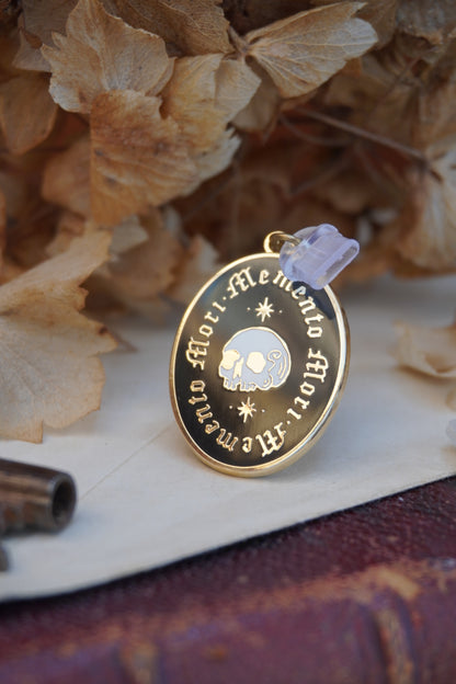 A close up of an oval enamel dust plug pendant black background, gold and white skull an stars, and the text 'Memento Mori' framed twice in gold, styled on vintage paper with dried flowers, a vintage key, and a crystal.
