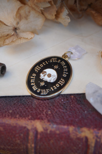 An oval enamel dust plug pendant black background, gold and white skull an stars, and the text 'Memento Mori' framed twice in gold, styled on vintage paper with dried flowers, a vintage key, and a crystal.