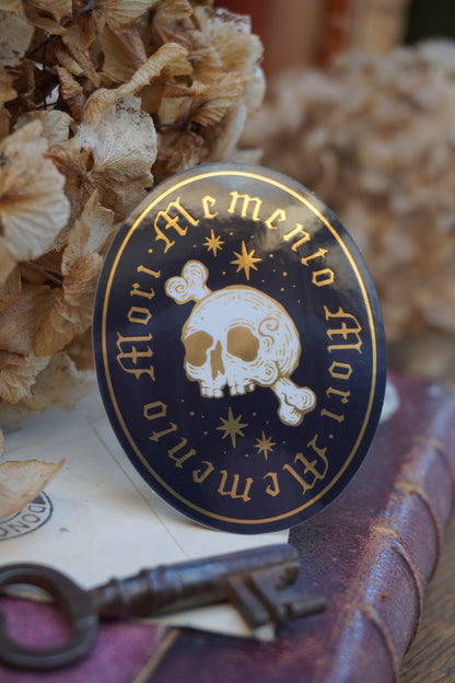 A close-up of an ovel sticker in white, black and gold, with gold lettering reading 'Memento Mori' and a white skull-and-crossbones motif, shown resting on an old book with dried flowers in the background and a vintage key in front