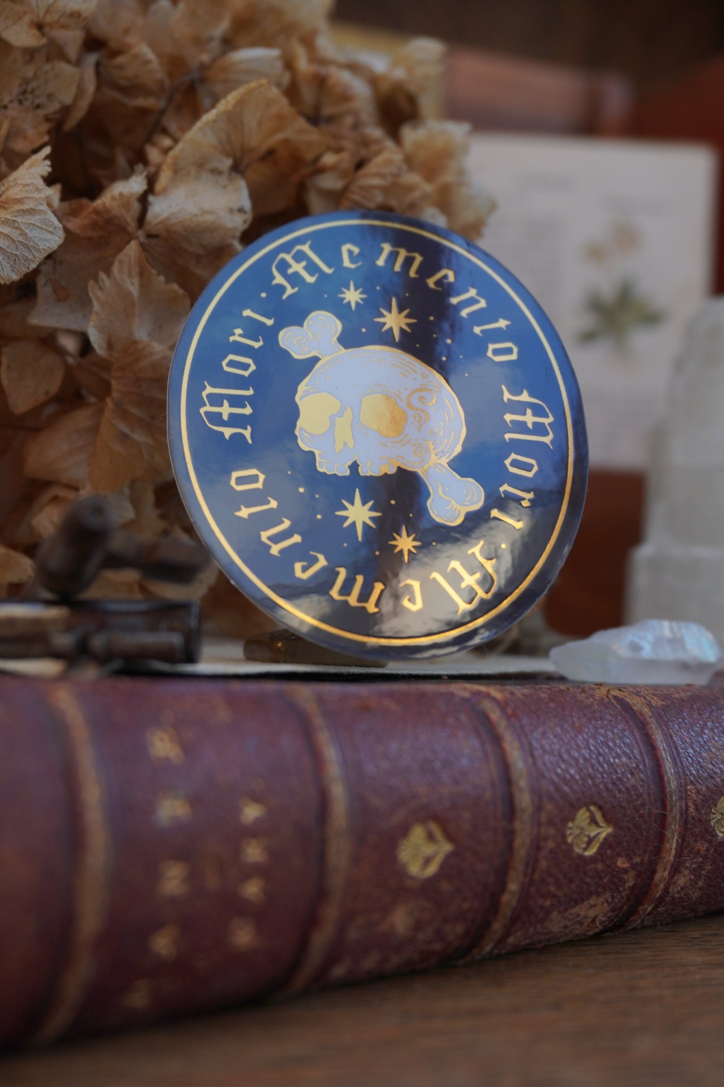 A close-up of an oval sticker in white, black and gold, with gold lettering reading 'Memento Mori' and a white skull-and-crossbones motif, shown resting on an old book with dried flowers in the background