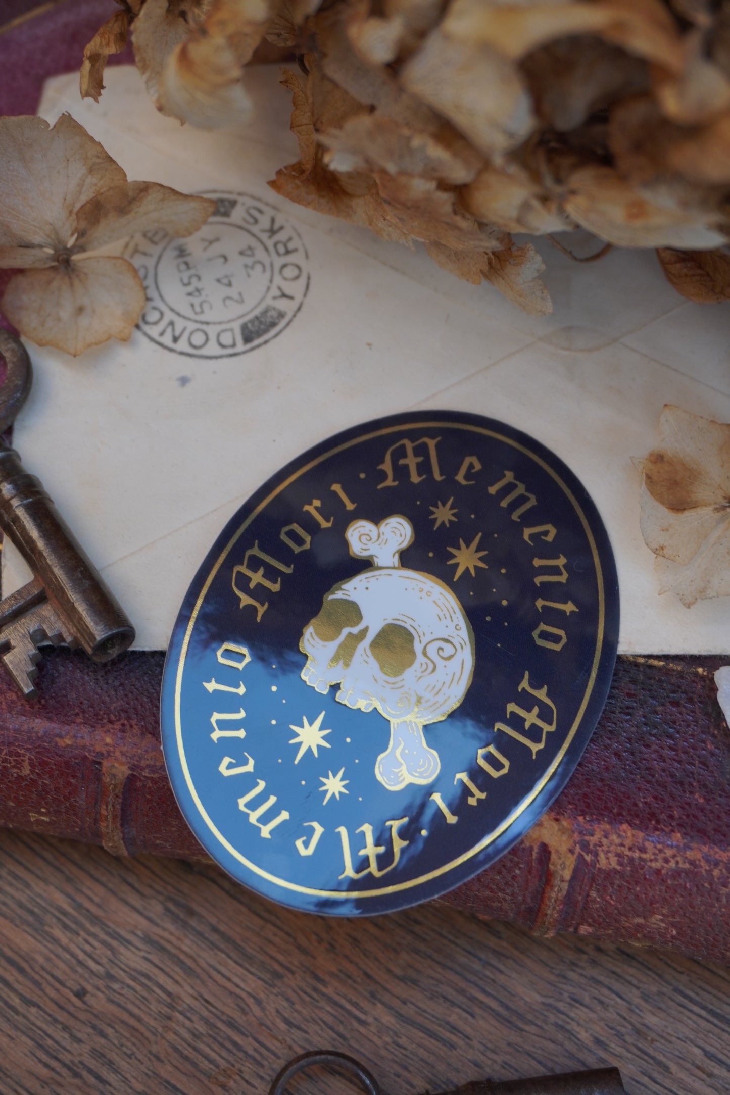 A close-up of an ovel sticker in white, black and gold, with gold lettering reading 'Memento Mori' and a white skull-and-crossbones motif, shown resting on an old book with dried flowers in the background