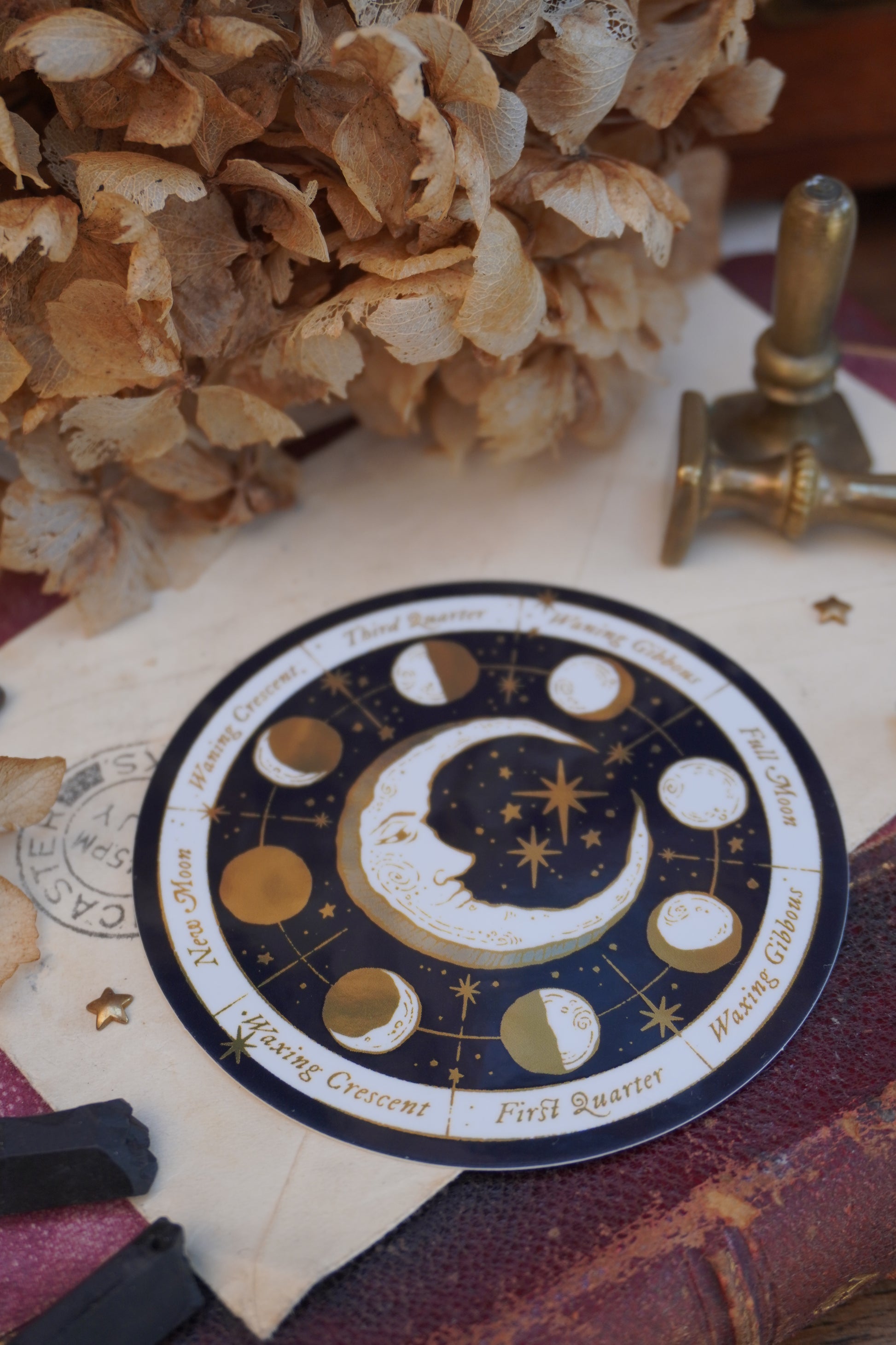 A circular moon phase calendar sticker with gold, cream and white celestial illustrations, and gold text labels, styled on a vintage book with dried flowers in the background and decorative objects