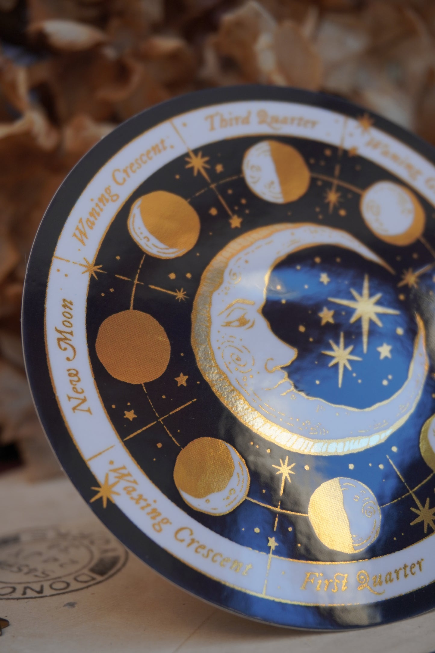Close up of a circular moon phase calendar sticker with gold, cream and white celestial illustrations, and gold text labels, styled on a vintage book with dried flowers in the background 