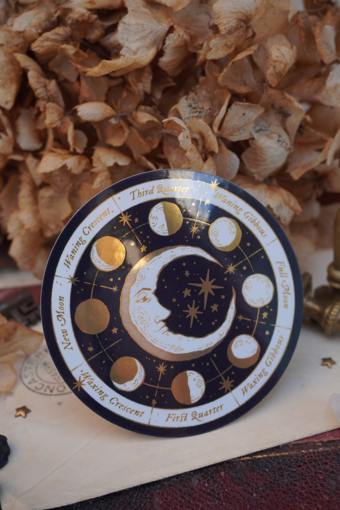 A circular moon phase calendar sticker with gold, cream and white celestial illustrations, and gold text labels, styled on a vintage book with flowers in the background.