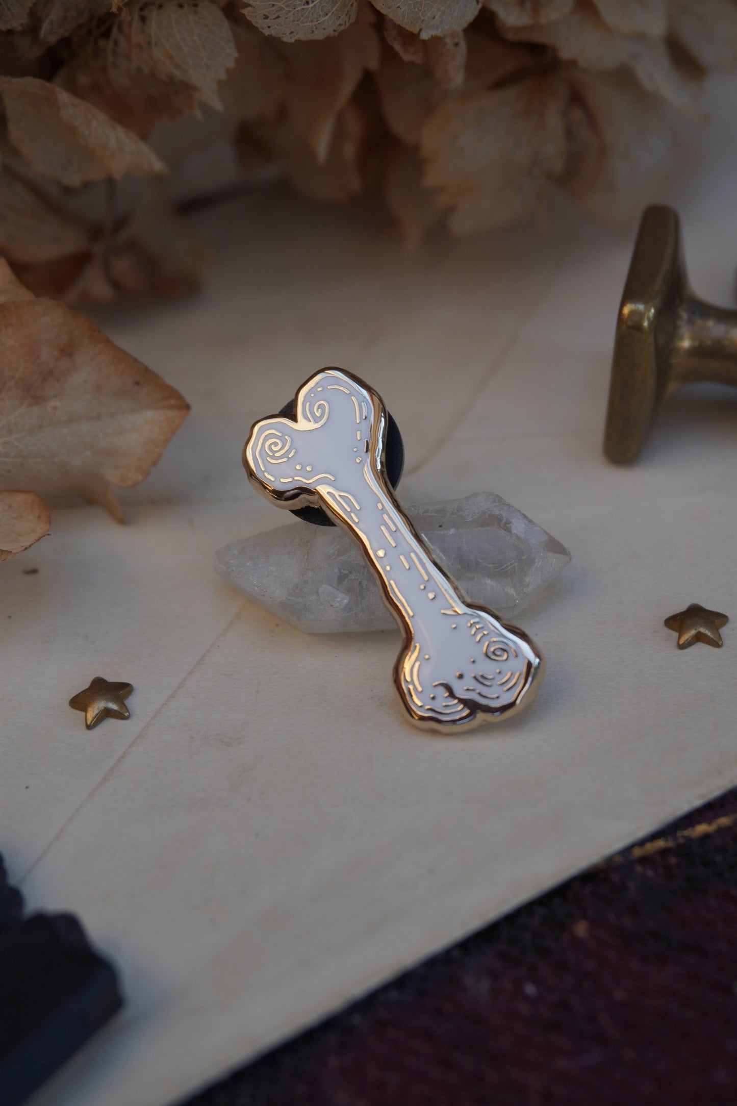 A bone shaped enamel pin in white and gold, resting on a small white crystal displayed on vintage paper on an old book with dried flowers and wax seals in the background