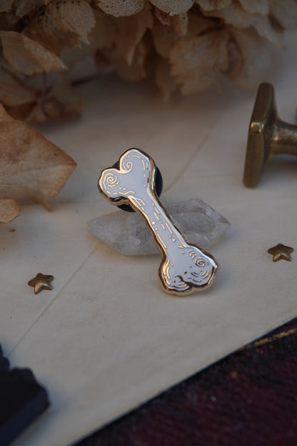 A bone shaped enamel pin in white and gold, resting on a small white crystal displayed on vintage paper on an old book with dried flowers and wax seals in the background